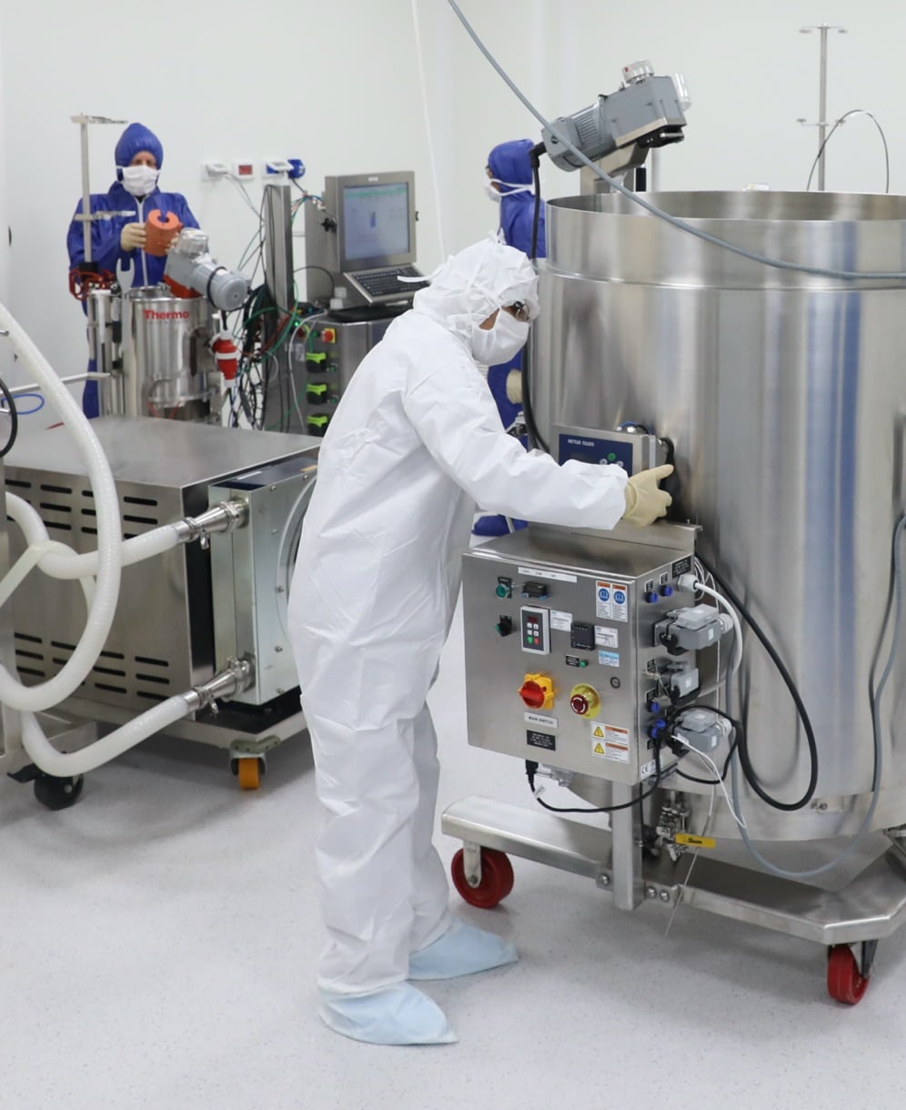 VBL production technicians in blue and white lab hoodies standing next to large stainless steel bioreactors in a clean room.